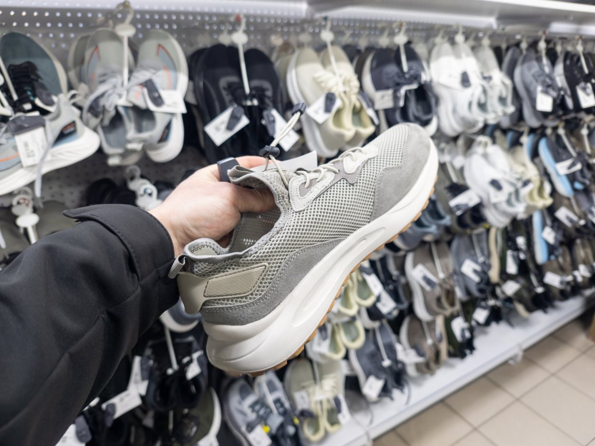 Hand holding a grey sneaker against shelves of footwear in a shoe store